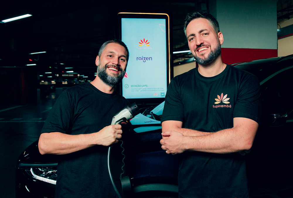 Imagem mostra dois homens de camisetas pretas, sorrindo e segurando plug de abastecimento de carros elétricos.