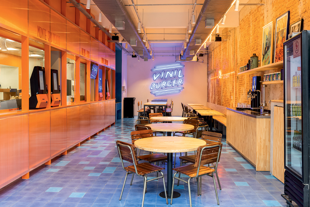 Ambiente de hamburgueria com totens de autoatendimento à esquerda, mesas no centro do salão e, ao fundo, o nome do estabelecimento escrito em letras de luz LED