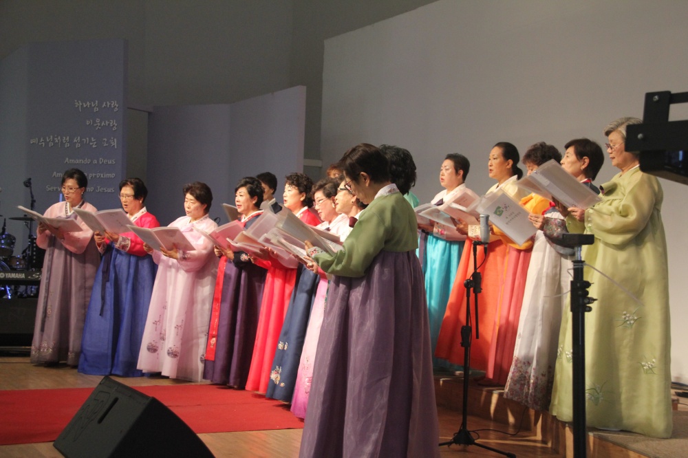 Foto mostra um coral de mães coreanas, que usam trajes longos tradicionais. Cada uma delas usa um traje de uma cor diferente.