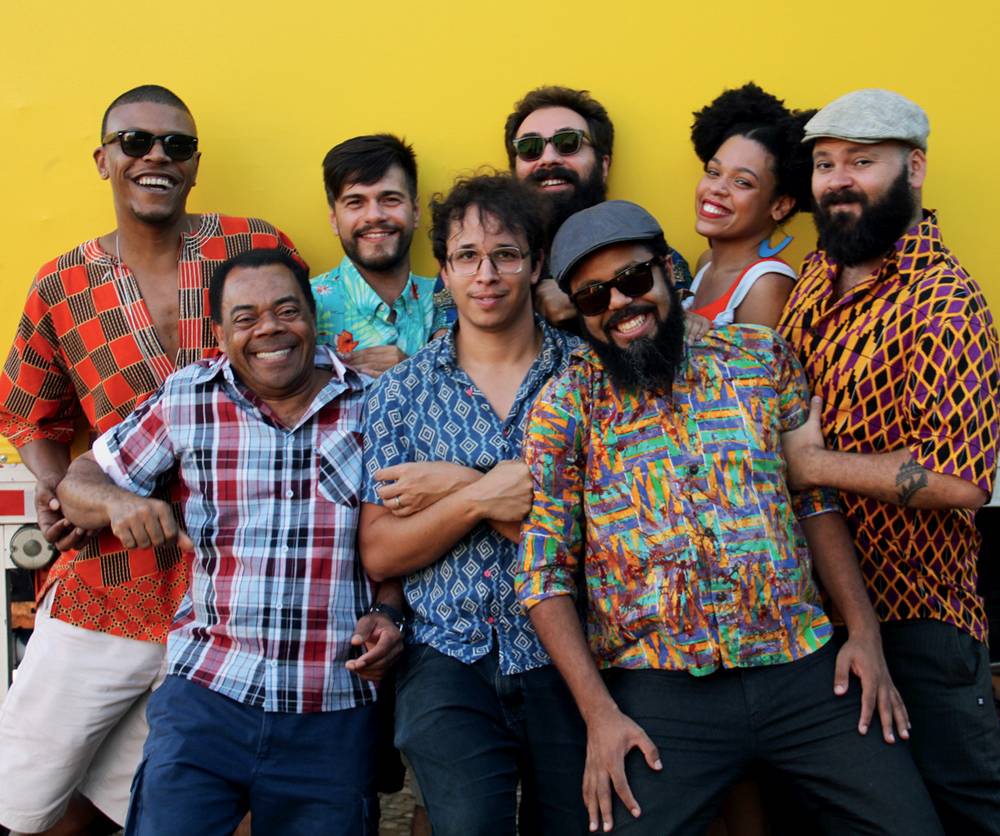 Sete homens e uma mulher vestem roupas coloridas e posam juntos sorrindo em frente a uma parede amarela. São um grupo musical.