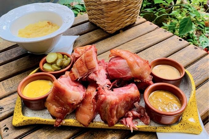 Sobre uma mesa de madeira, tábua de cerâmica de cor amarela serve itens como joelho de porco, mostardas e picles fatiado