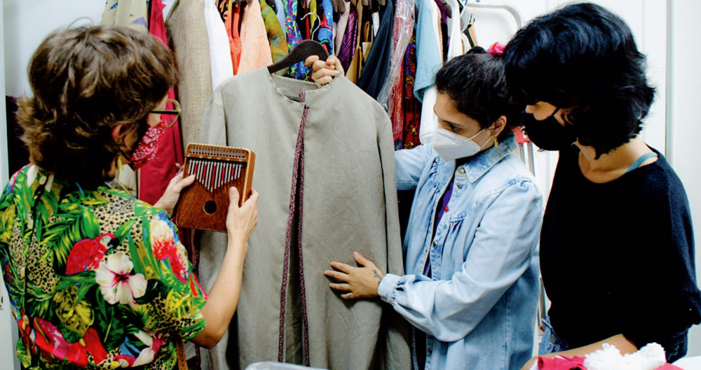 Uma mulher segura um blazer cinza, enquanto outra olha para ela. Uma terceira mulher segura uma kalimba (instrumento africano) próxima à roupa. Todas usam máscara. Elas estão em um ateliê.