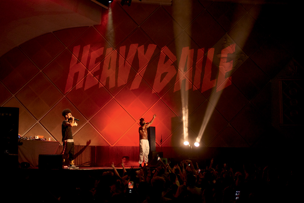 Imagem mostra pessoas em frente a palco iluminado por luzes brancas e vermelhas. Dois homens com microfones, um deles sem camisa, estão em cima do palco. Na parede do espaço, luzes vermelhas projetam o nome "Heavy Baile".