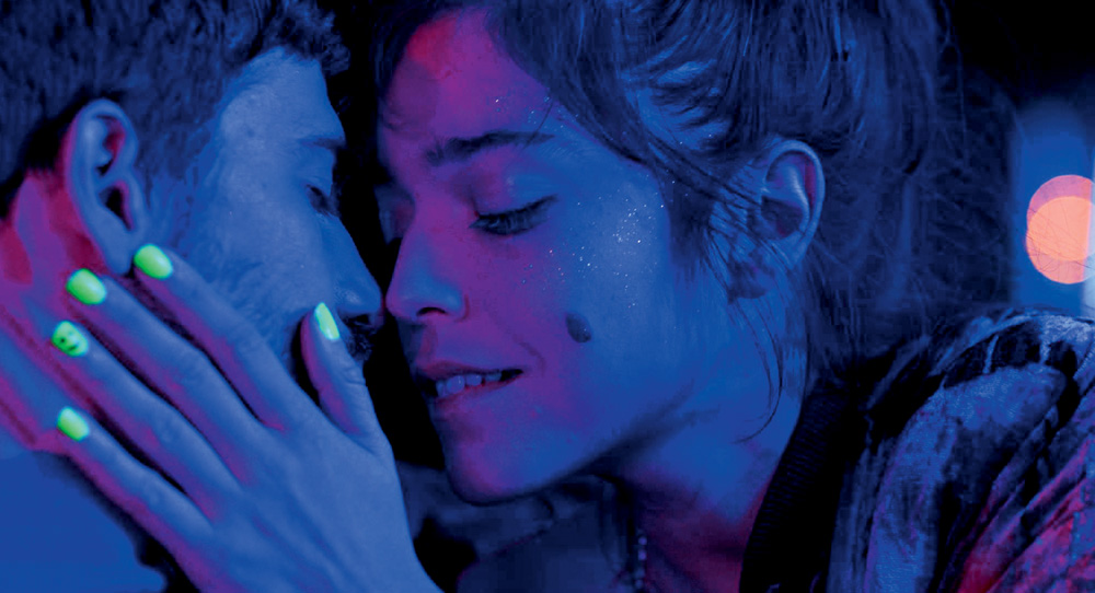 Imagem mostra casal se beijando sob luz azul intensa. A mulher, à direita, está com a mão no rosto do homem, à esquerda. Ela tem unhas com esmalte luminescente amarelo.