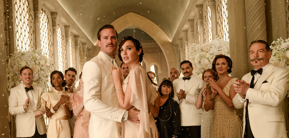 Imagem mostra casal abraçado entre círculo de pessoas, todos com trajes de gala brancos. O casal tem olhar preocupado.
