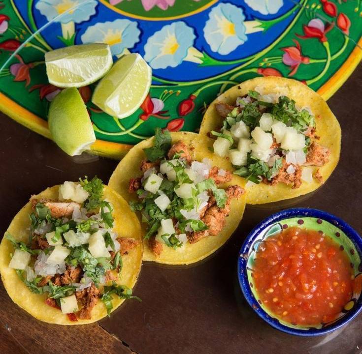 Três tacos abertos e recheados em cima de uma mesa de madeira. Ao lado há um pote colorido com molho.