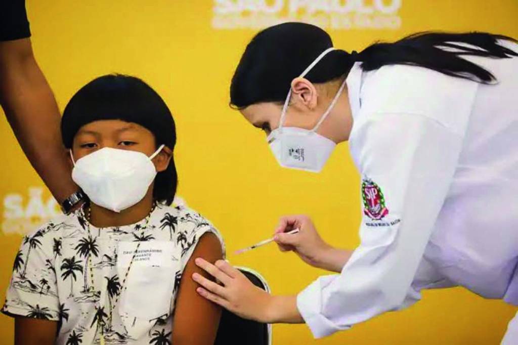 Menino indígena usa uma máscara branca e é vacinado por uma enfermeira de jaleco e máscara brancos