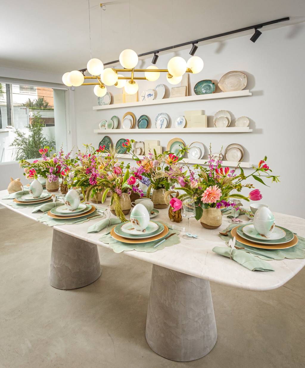Showroom Casa Costa: foto exibe mesa branca repleta de flores rosas e pratos de porcelana. Ao fundo, prateleiras brancas com pratos expostos.