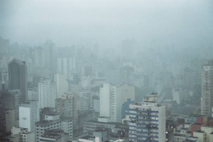Imagem mostra horizonte de prédios sob céu nublado com chuva fina.