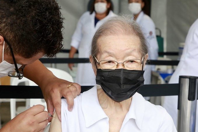 Imagem mostra mulher idosa de máscara e óculos. Ela está sentada e recebe uma injeção no braço