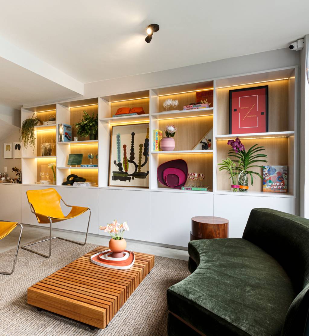 Imagem exibe sala de estar com mesa de centro baixa, prateleiras iluminadas, sofá preto e duas cadeiras amarelas.