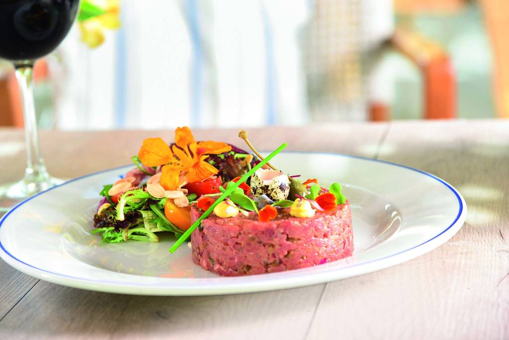 Prato raso de louça branca é usado para servir steak tartare com salada decorada por flores comestíveis