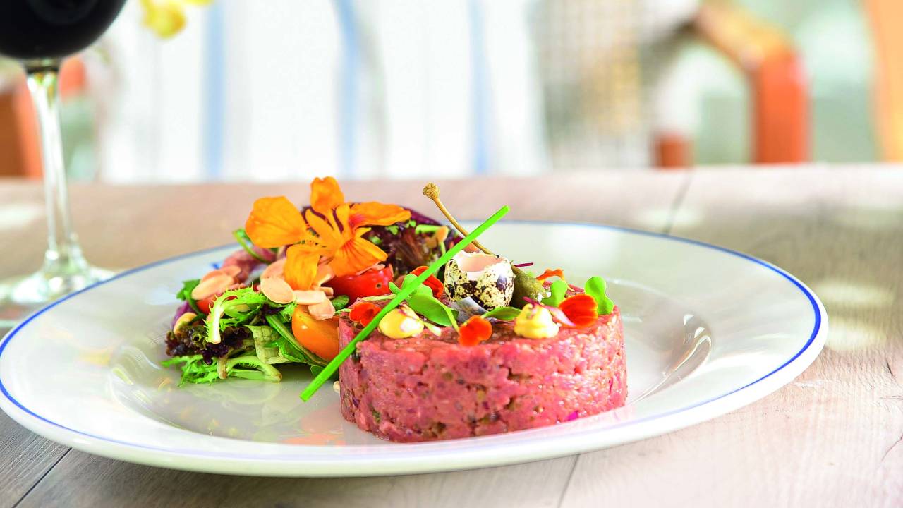 Prato raso de louça branca é usado para servir steak tartare com salada decorada por flores comestíveis