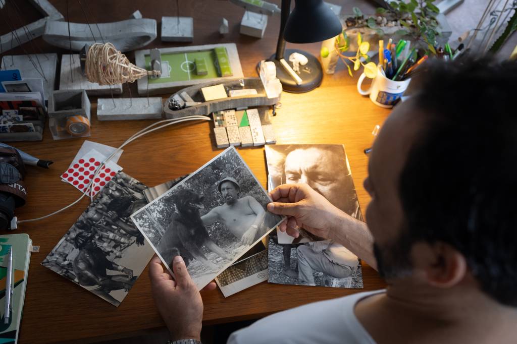 Imagem mostra Noel sentado em mesa, segurando fotos do pai, Orlando