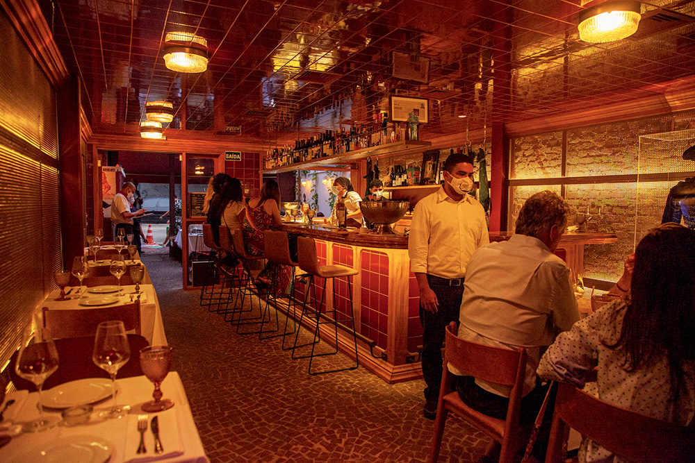 Ambiente de restaurante de paredes decoradas por azulejos de cor avermelhada e iluminado por luzes amareladas. No balcão do bar, há garrafas expostas e banquetas altas que acomodam os clientes.