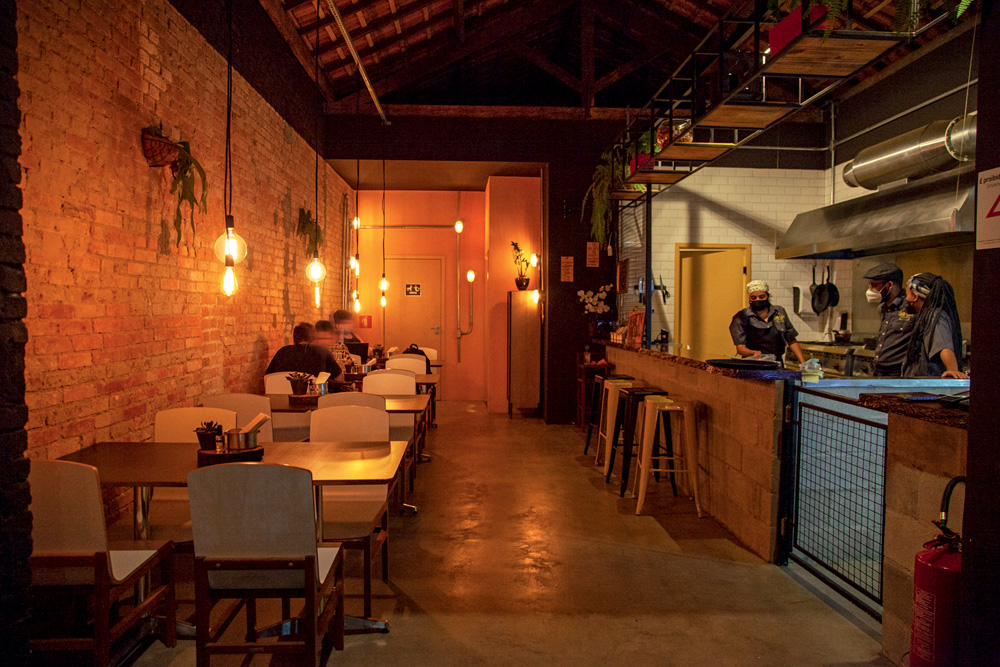 Ambiente do bar Lapa Lapa decorado por lâmpadas penduradas, mesas e cadeiras de madeiras, um balcão com banquetas altas e uma cozinha visível