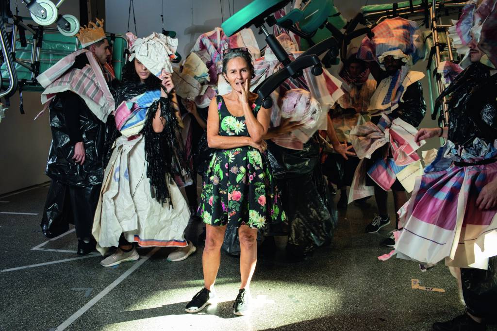 Bia Lessa Imagem mostra mulher com vestido estampado em pé me meio a diversas pessoas vestindo figurinos coloridos.