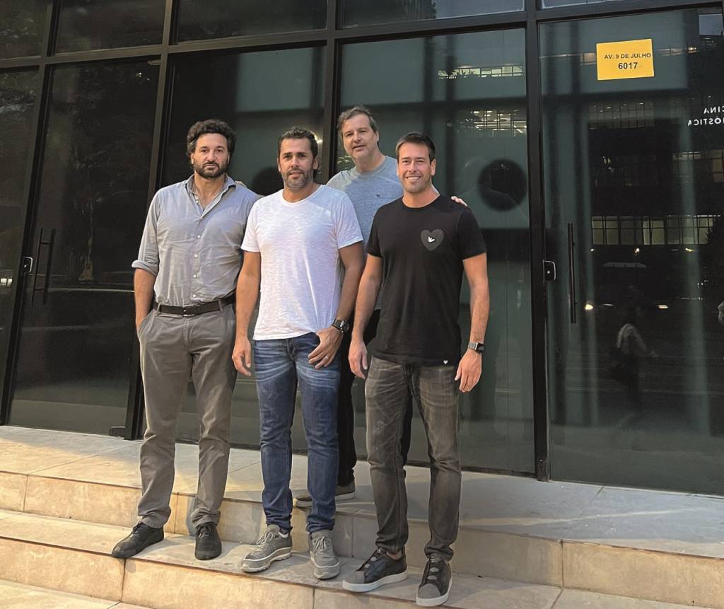 Rico, Tiago, Gutti e Marcos (a partir da esquerda) posam em escada na entrada de balada. Todos vestem calças escuras, o primeiro camisa clara e os demais camisetas.