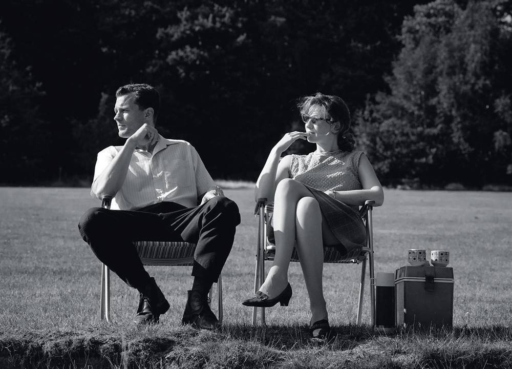 Imagem em preto e branco mostra homem e mulher sentados em cadeiras colocadas em gramado.