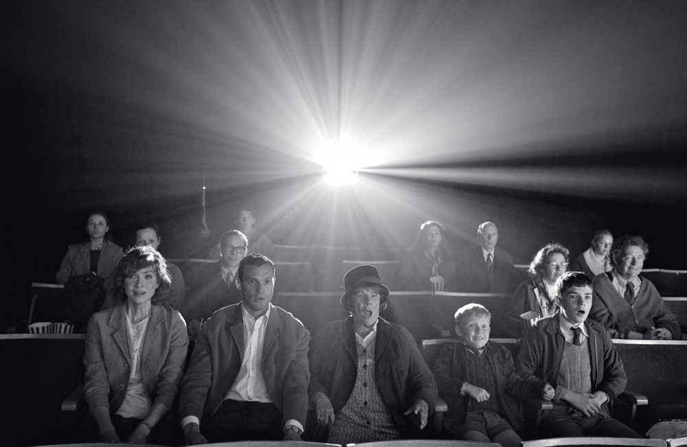 Imagem em preto e branco mostra plateia de cinema sentada. Ao fundo, a luz do projetor.