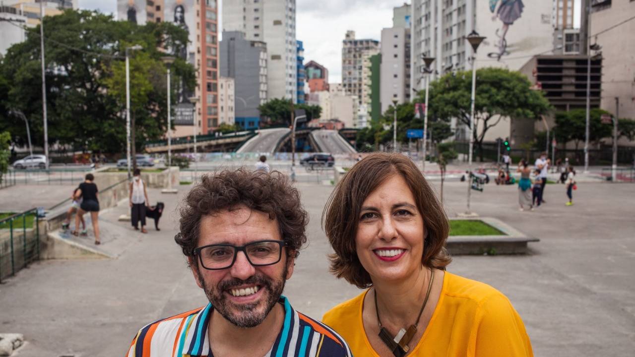 Clayton Melo e Denize Bacoccina posam com a Roosevelt de fundo. Ele usa camisa listrada colorida e ela uma camiseta amarela.