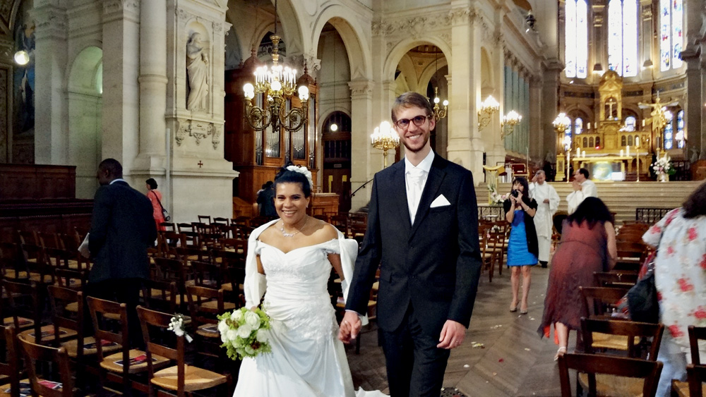 Luciane e Paul no dia de seu casamento. Ele veste um terno preto e óculos. Ela veste um longo vestido bordado com uma coroa. O buquê que ela segura tem rosas brancas.