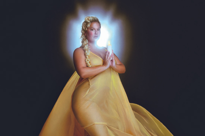 Imagem mostra mulher de vestido longo amarelo segurando uma vela com as mãos.