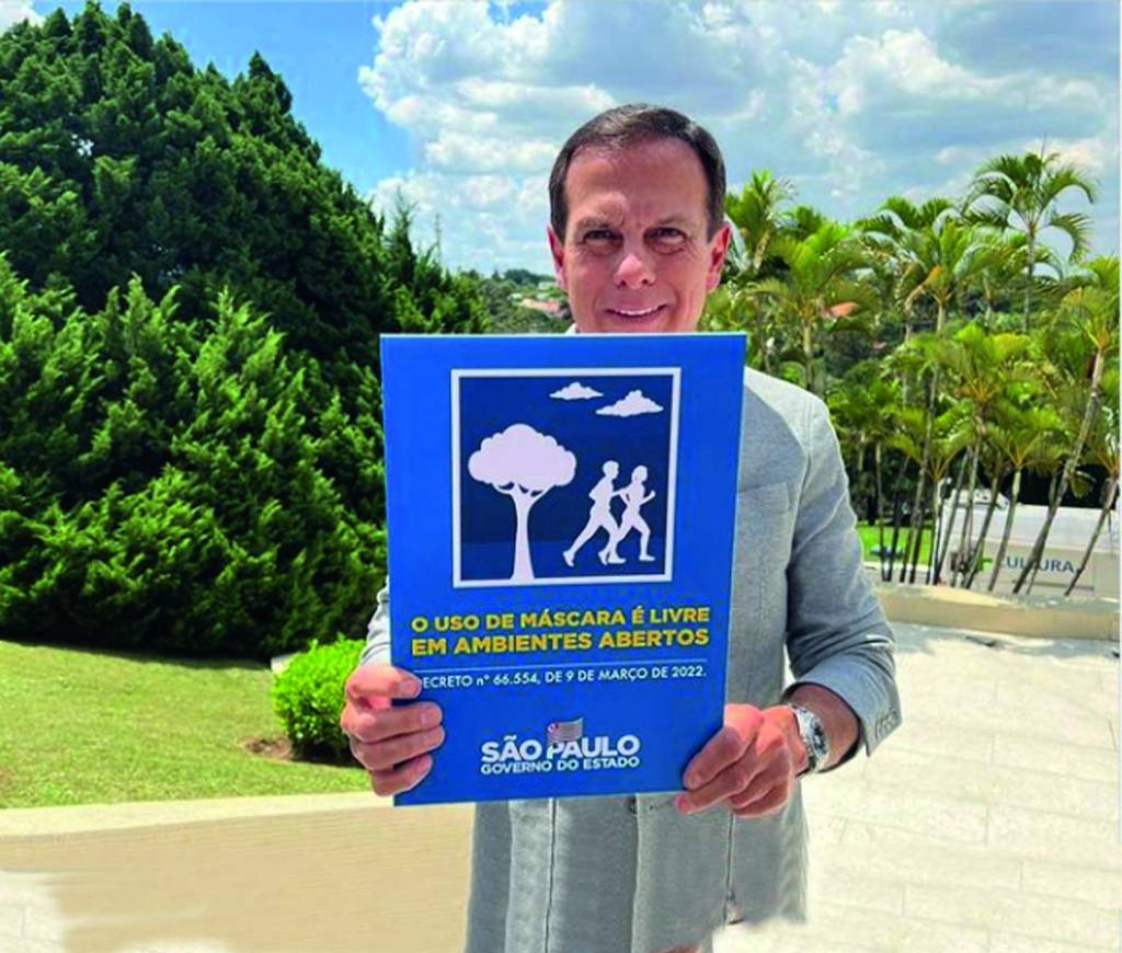 O governador João Doria aparece ao ar livre, sem máscara, segurando um cartaz azul sobre a nova medida