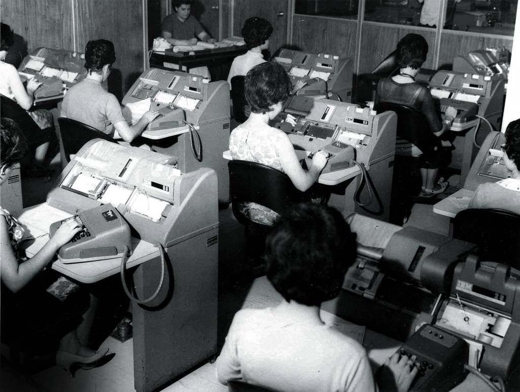 Imagem em preto e branco mostra diversas mulheres sentadas em escrivaninhas trabalhando em máquinas de escrever