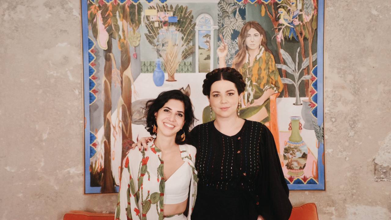 Fernanda Feher e Betina de Luca posam lado a lado, com obra colorida ao fundo. A da esquerda tem cabelos escuros e curtos, a da direita está com os cabelos presos.