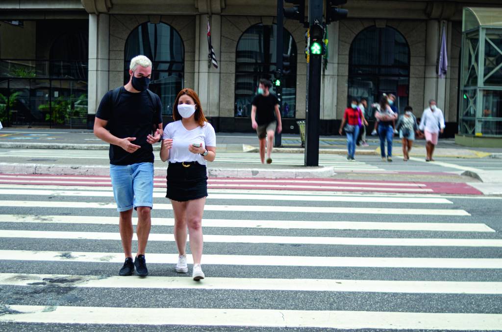 Um homem e uma mulher andam por um cruzamento na Avenida Paulista. Ambos usam máscaras
