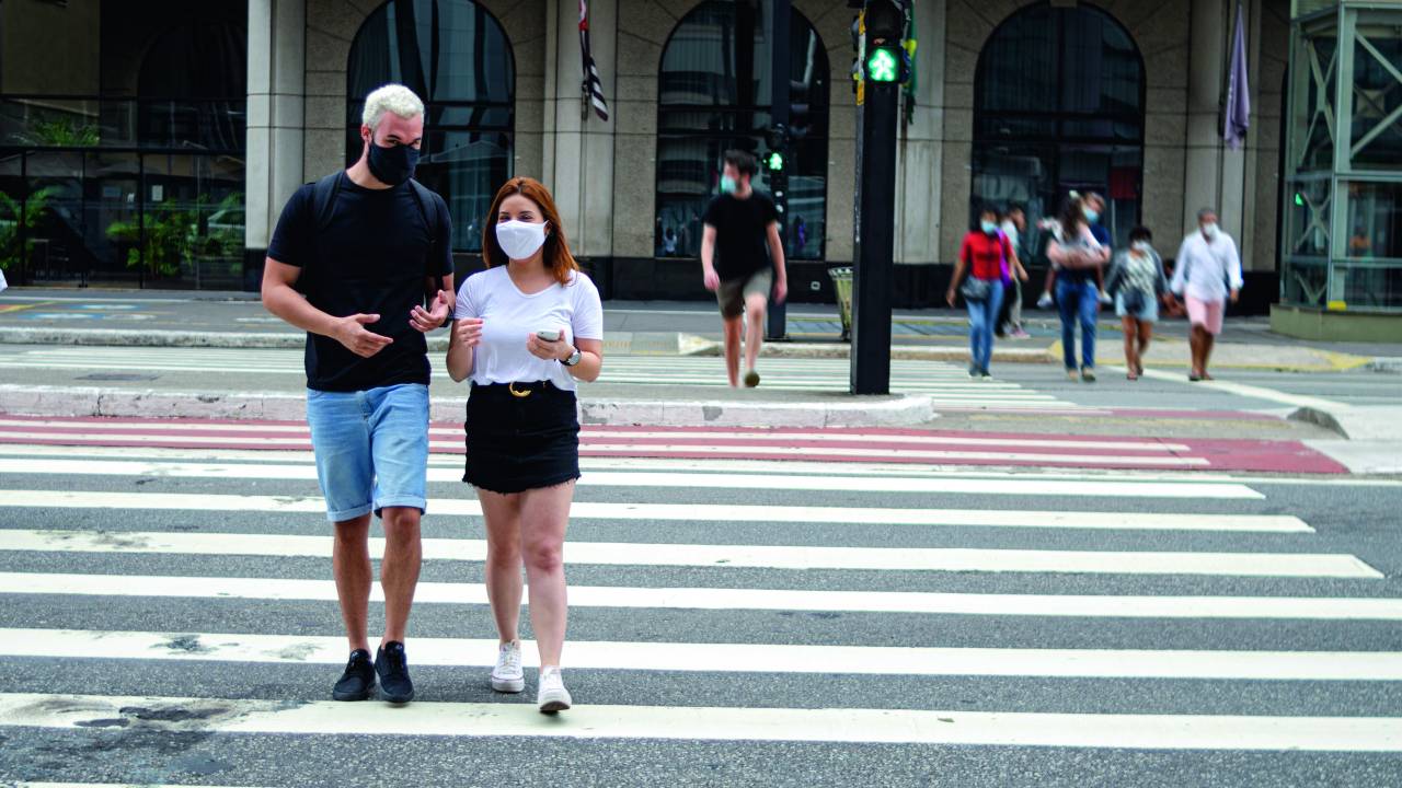 Um homem e uma mulher andam por um cruzamento na Avenida Paulista. Ambos usam máscaras