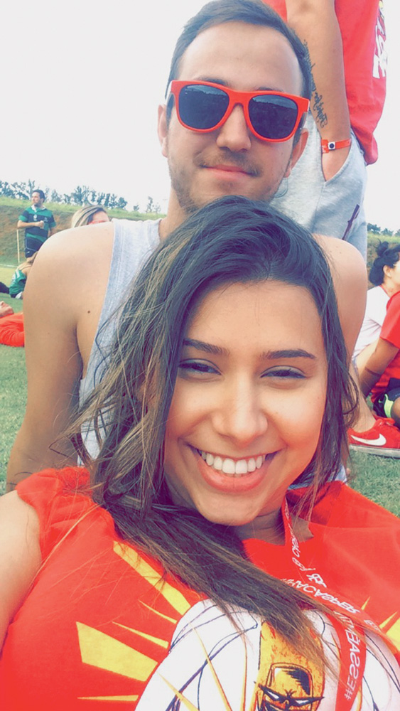 Beatriz faz uma selfie com Felipe atrás. Ele usa um óculos de sol e ela uma blusa vermelha. Eles estão sentados em um campo gramado