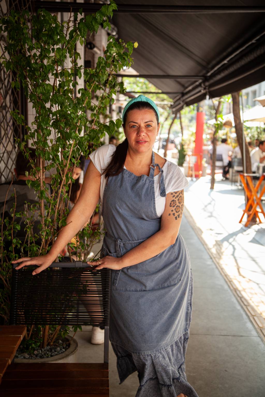 A chef Janaína Rueda de avental apoiada em cadeira de madeira na calçada d'A Casa do Porco.