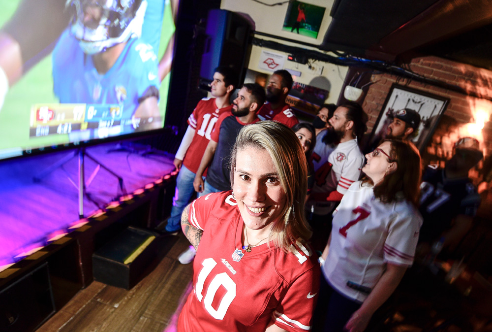 Imagem mostra mulher loira vestindo camiseta de time vermelha em meio a aglomeração de pessoas em bar.