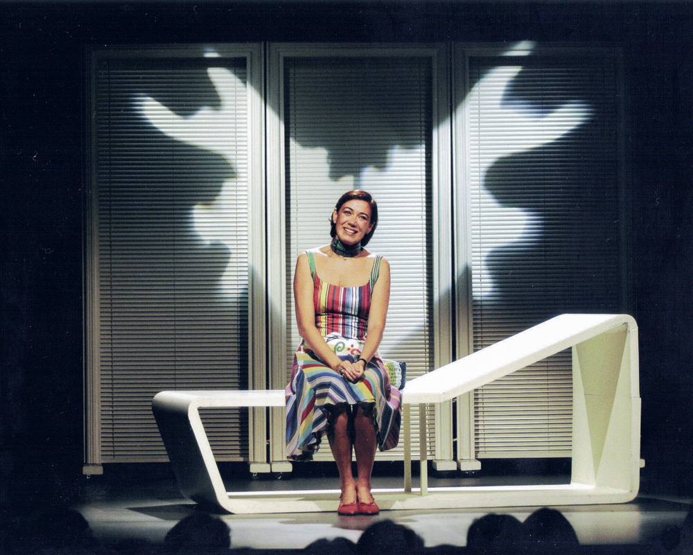 Lilia usa um vestido listrado colorido e está sentada em um banco comprido branco no meio do palco.