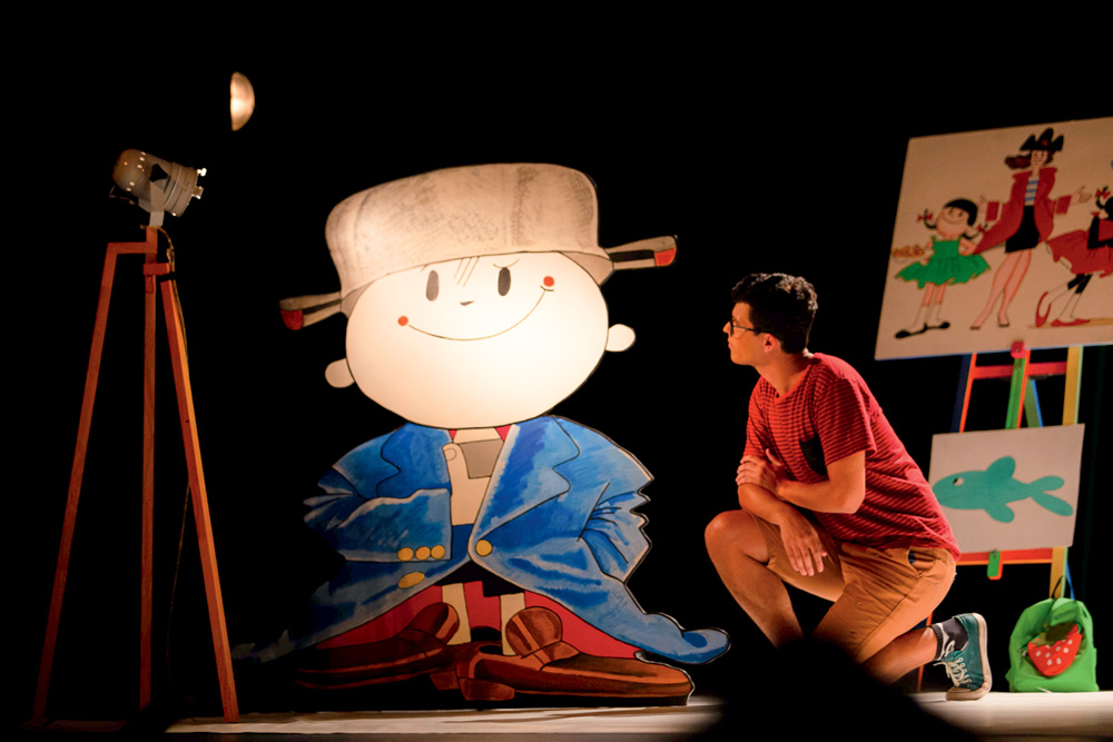 Um ator interpreta um dos netos de Ziraldo e se posiciona agachado ao lado do cartaz do Menino Maluquinho.