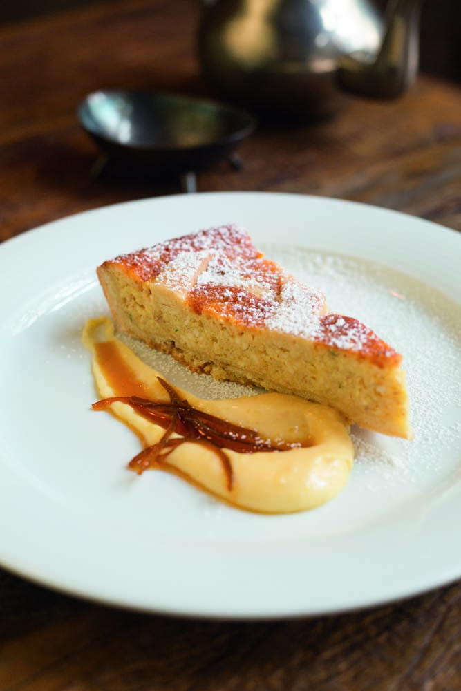 Fatia de torta servida sobre pequeno prato raso de louça branca e junto de creme de cor levemente amarelada