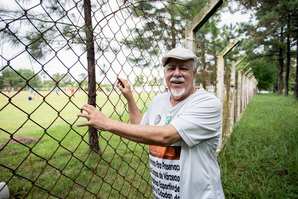 Senhor negro sorri ao lado da grade de um campo de futebol gramado