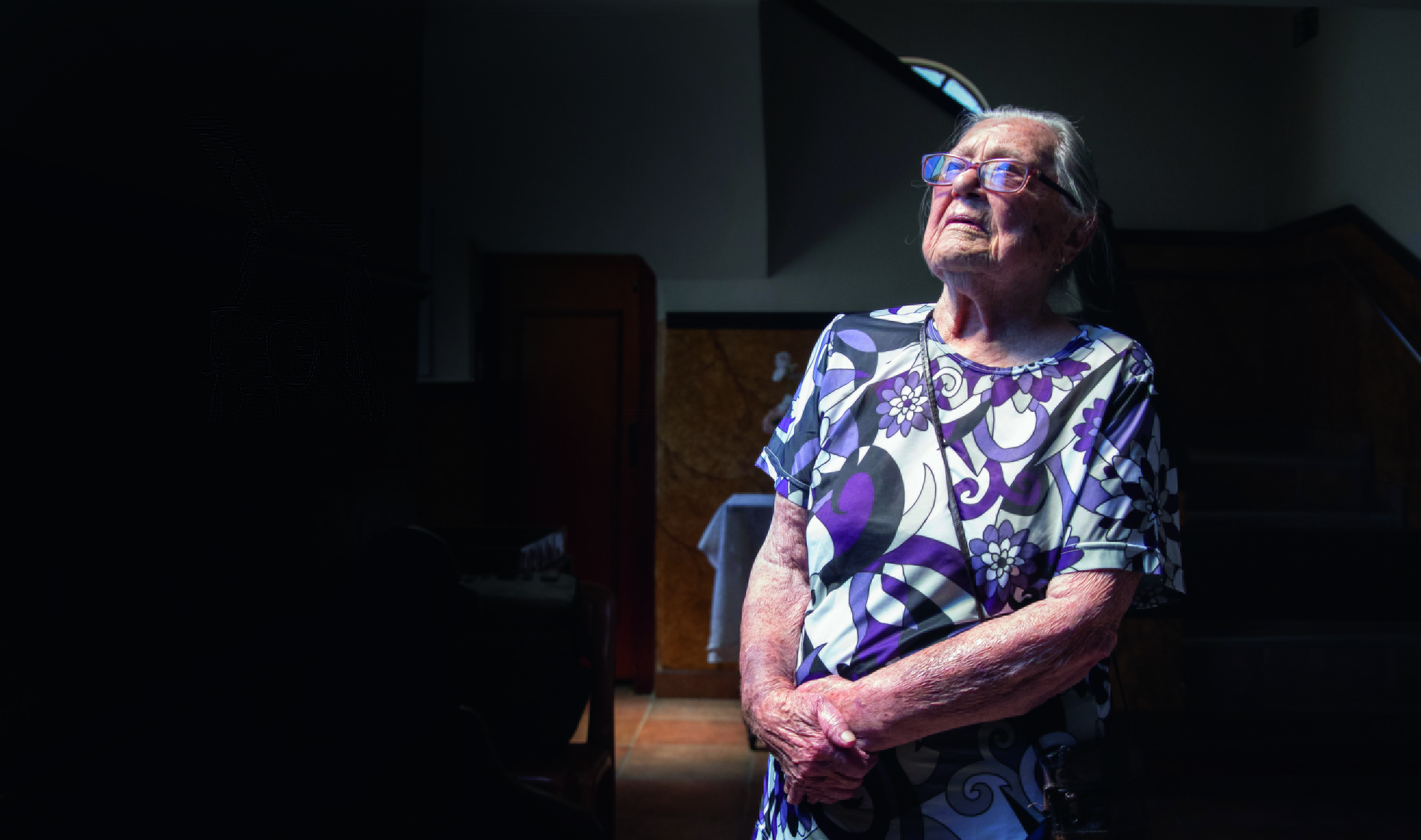 Imagem mostra mulher idosa com os braços cruzados sobre a barriga, em foto escura.
