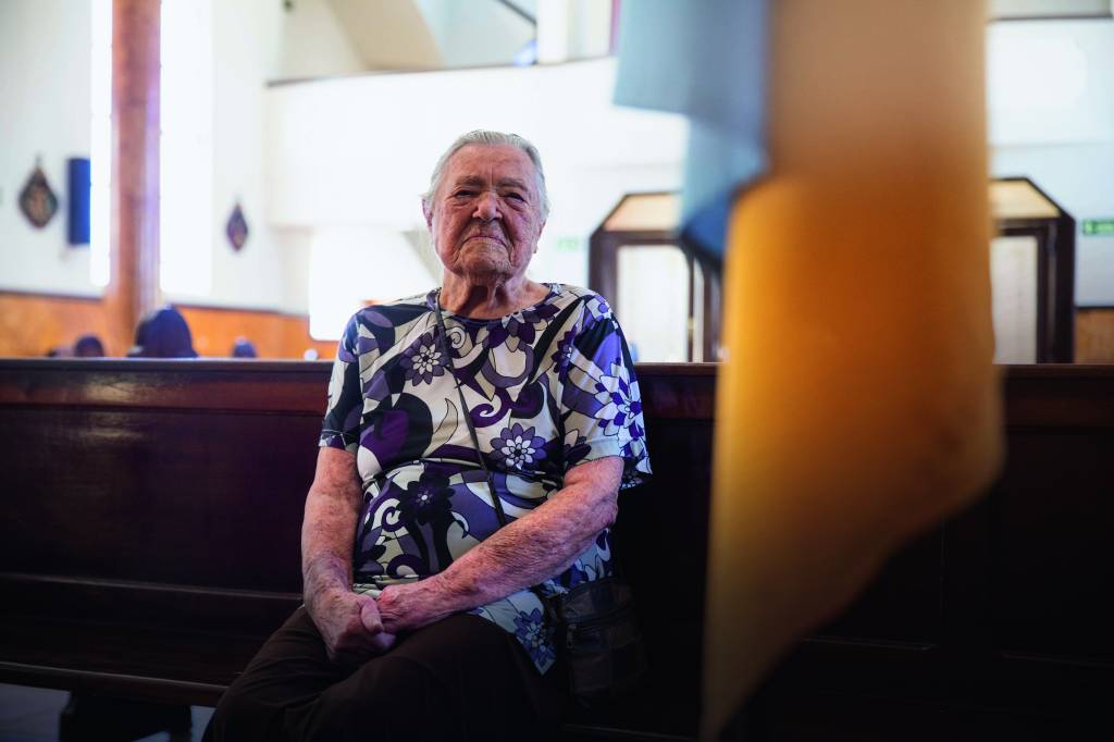 Imagem mostra mulher idosa sentada em banco de igreja.