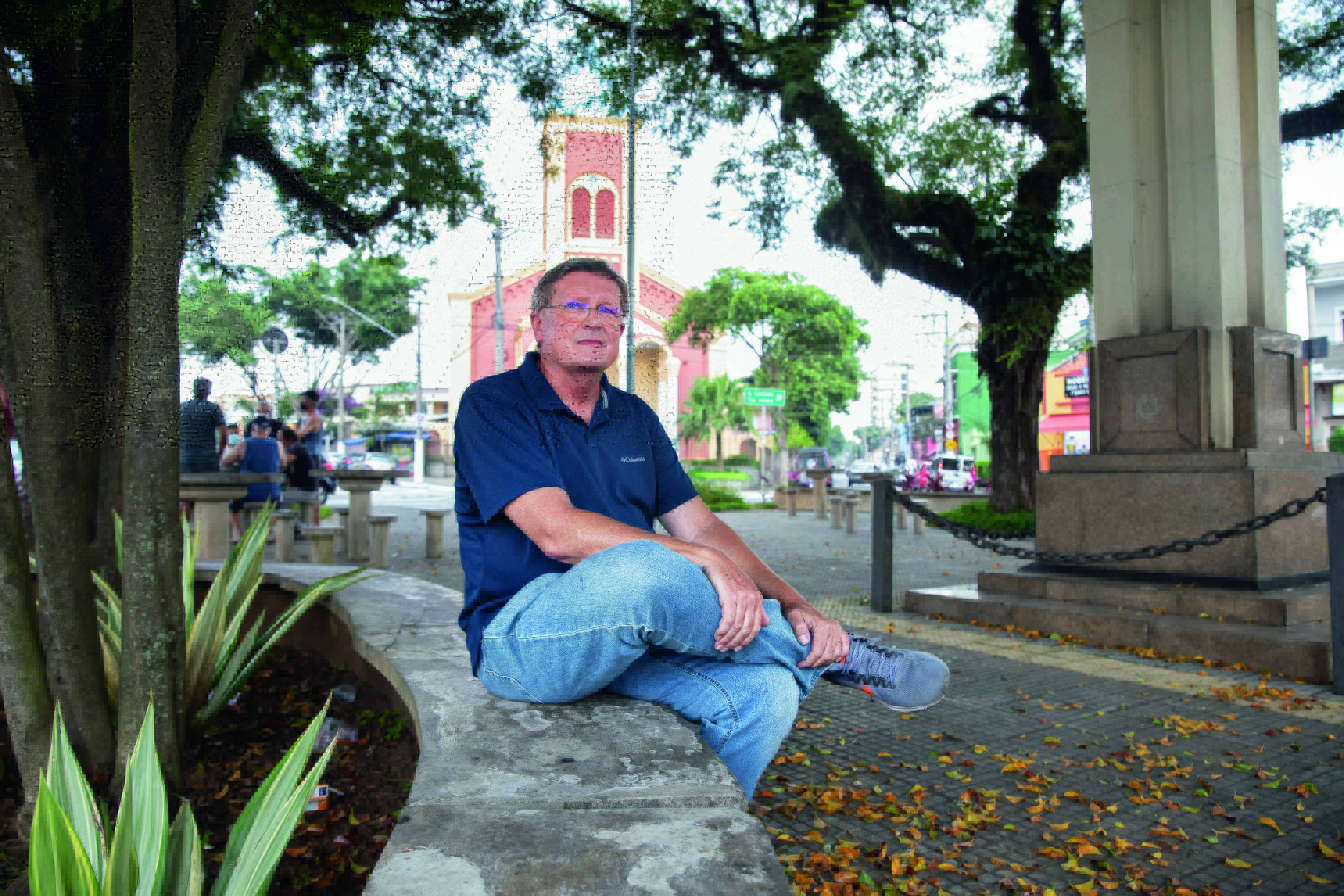 Imagem mostra homem de calça jeans e camisa polo azul marinho sentado em praça. Ao fundo, a fachada de uma igreja.