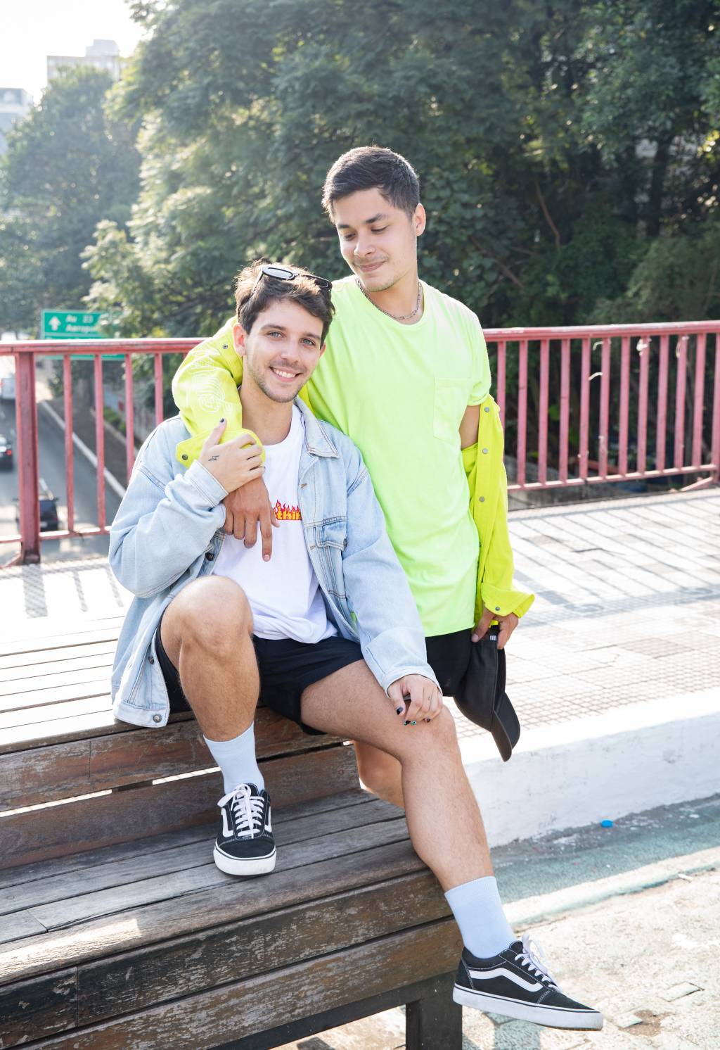 Victor e Vinicius lado a lado em rua da Liberdade. Victor veste camiseta branca com jaqueta jeans clara e Vinicius veste camiseta e jaqueta na cor amarelo neon. Vinicius está de pé e olha para Victor, sentado em banco na ponte da Liberdade.