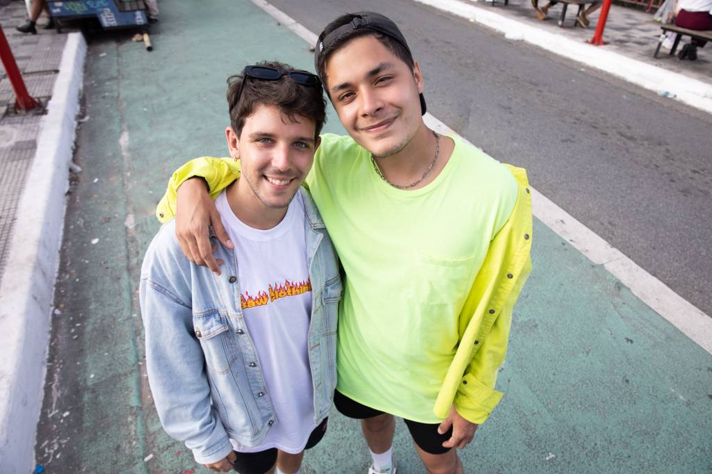 Victor e Vinicius lado a lado em rua da Liberdade. Victor veste camiseta branca com jaqueta jeans clara e Vinicius veste camiseta e jaqueta na cor amarelo neon.