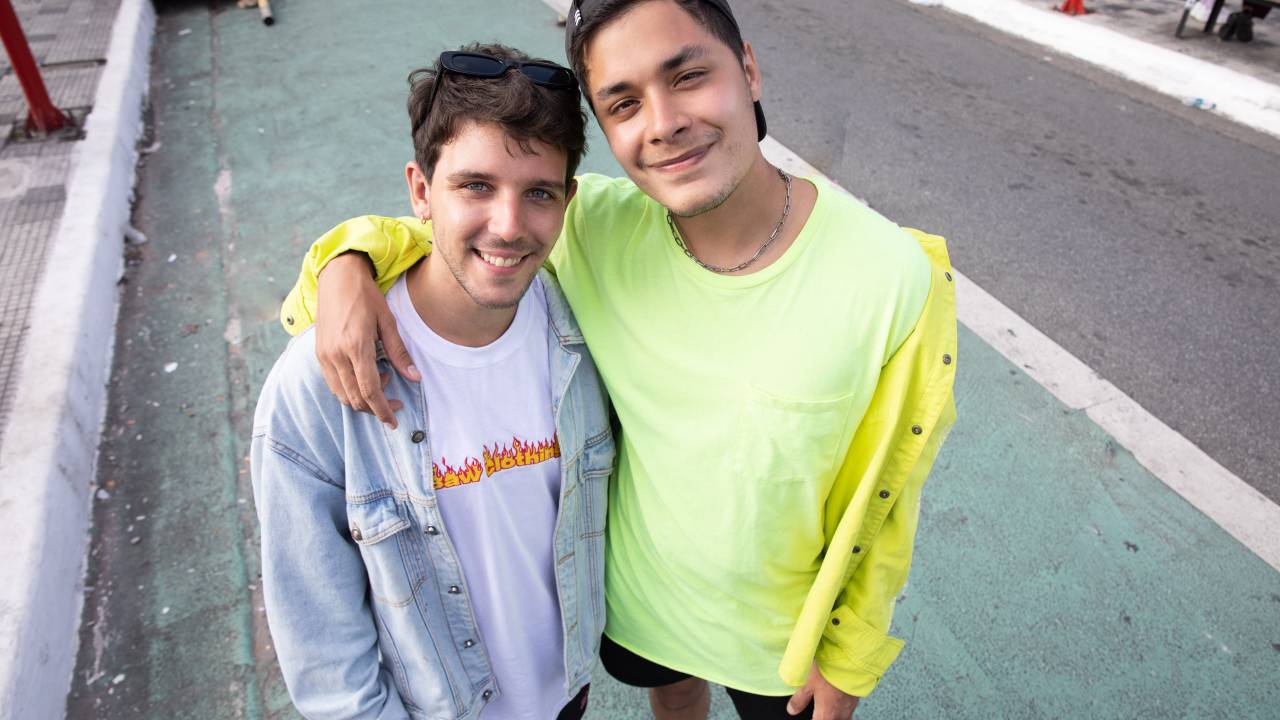 Victor e Vinicius lado a lado em rua da Liberdade. Victor veste camiseta branca com jaqueta jeans clara e Vinicius veste camiseta e jaqueta na cor amarelo neon.