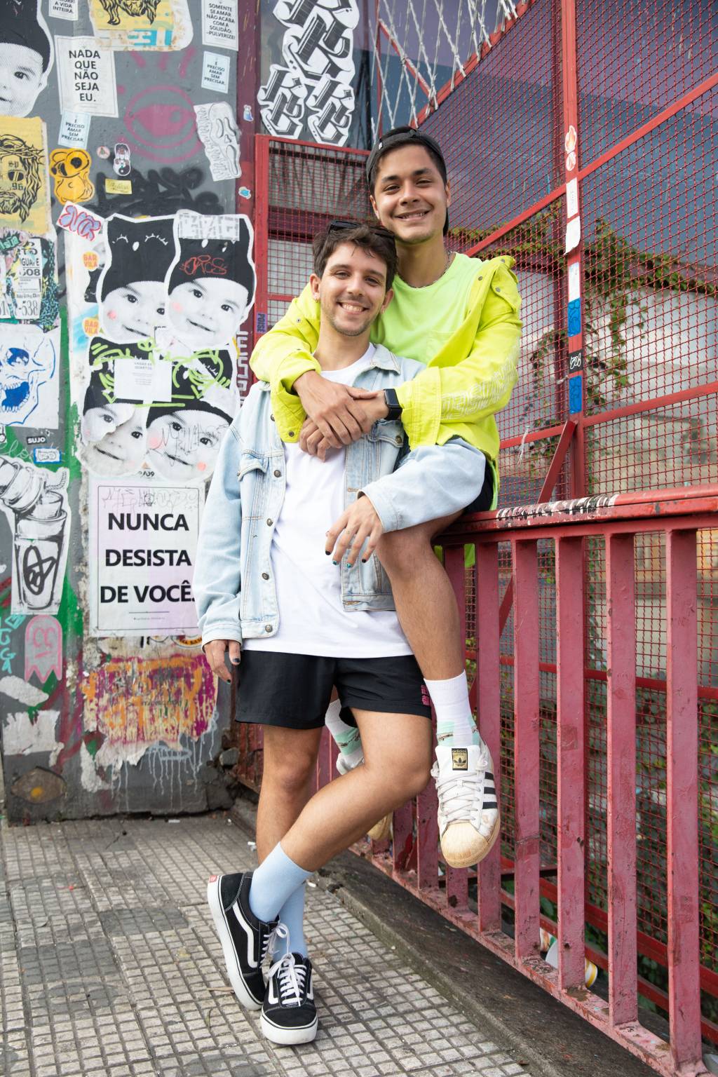 NOSSO LOUCO AMOR – Vinicius Lasserre e Victor Delmas (2) Casal posa encostado em grade vermelha de ponte da Liberdade. Victor está de pé e Vinicius está sentado na grade, apoiado no pescoço de Victor. Victor veste shorts preto, camiseta branca e jaqueta jeans clara. Vinicius veste tênis branco surrado e camiseta e jaqueta amarelo neon.