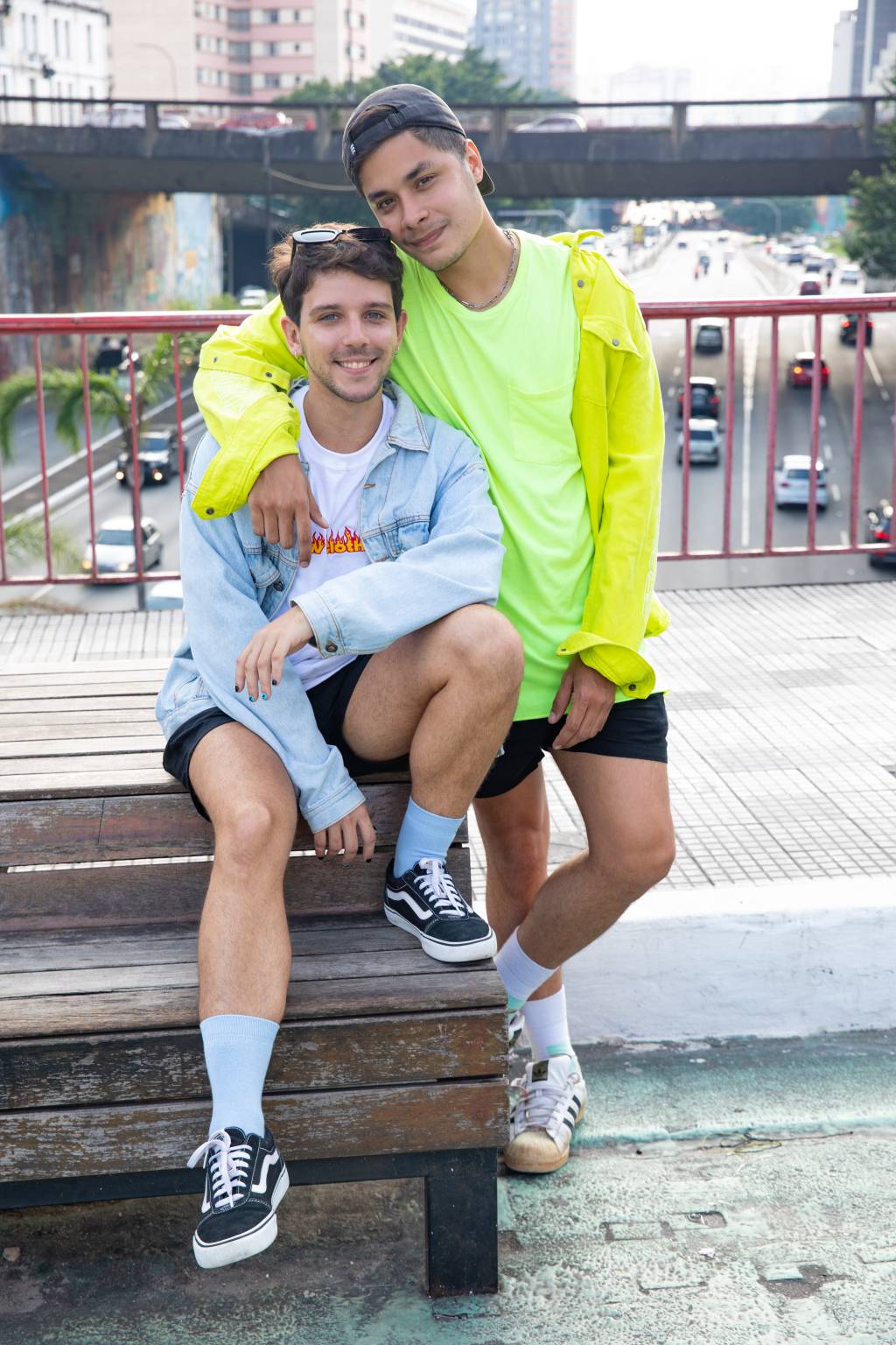 Victor e Vinicius lado a lado em rua da Liberdade. Victor veste camiseta branca com jaqueta jeans clara e Vinicius veste camiseta e jaqueta na cor amarelo neon. Vinicius está de pé e Victor sentado em banco na ponte da Liberdade.