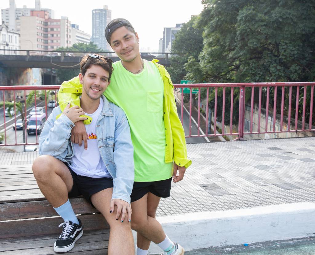 Victor e Vinicius lado a lado em rua da Liberdade. Victor veste camiseta branca com jaqueta jeans clara e Vinicius veste camiseta e jaqueta na cor amarelo neon. Vinicius está de pé e Victor sentado em banco na ponte da Liberdade.