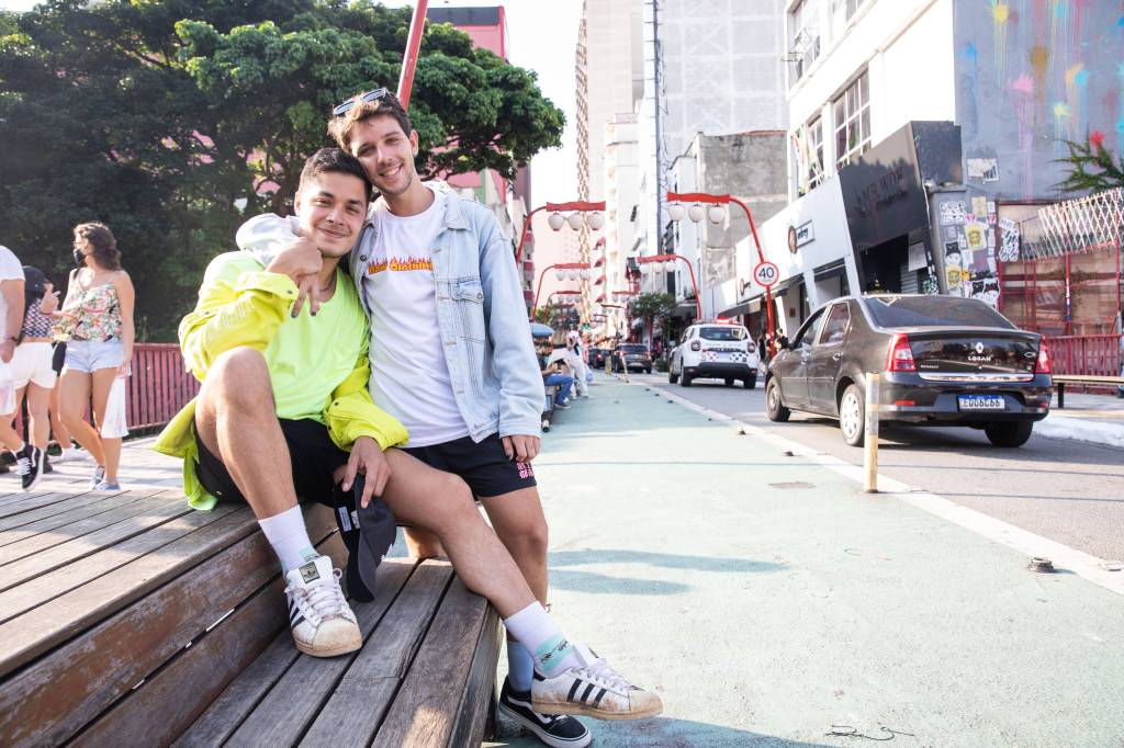 NOSSO LOUCO AMOR – Vinicius Lasserre e Victor Delmas (6) Victor e Vinicius lado a lado em rua da Liberdade. Victor veste camiseta branca com jaqueta jeans clara e Vinicius veste camiseta e jaqueta na cor amarelo neon. Victor encosta a cabeça em Vinicius, sentado em banco na ponte da Liberdade.