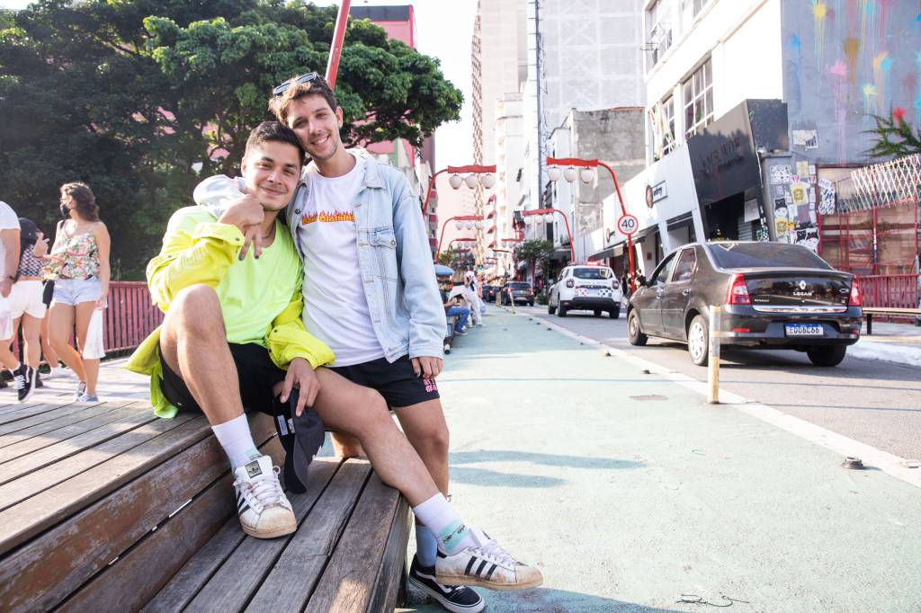 Victor e Vinicius lado a lado em rua da Liberdade. Victor veste camiseta branca com jaqueta jeans clara e Vinicius veste camiseta e jaqueta na cor amarelo neon. Victor encosta a cabeça em Vinicius, sentado em banco na ponte da Liberdade.